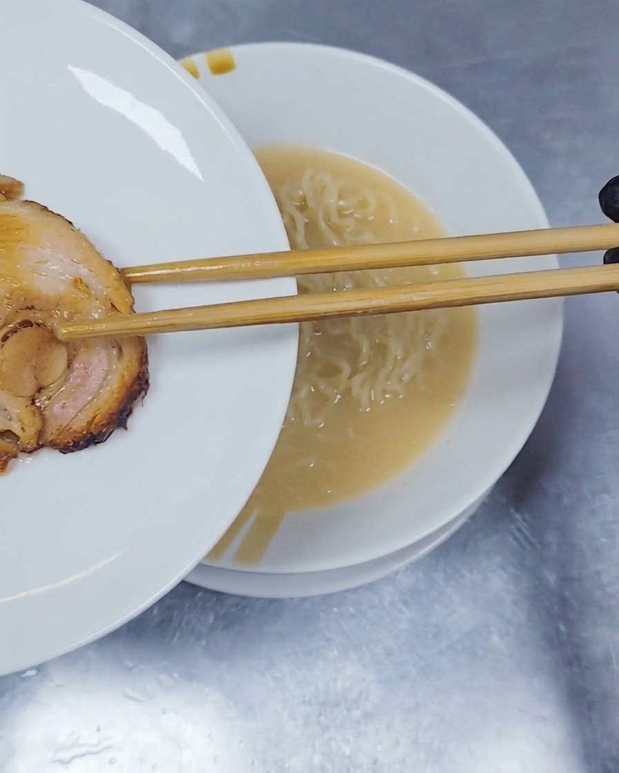 Eine Scheibe gerollter und geschmorter Chashu-Schweinebauch wird mit Holzstäbchen auf die Ramen-Nudeln gelegt.
