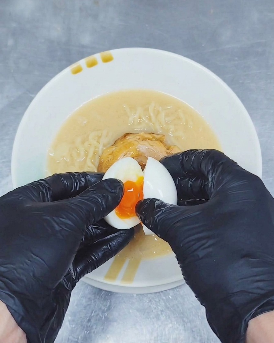 Ein Paar Hände in Handschuhen teilt ein weichgekochtes Ei über einer Schüssel Ramen, um ein flüssiges, goldenes Eigelb freizulegen.