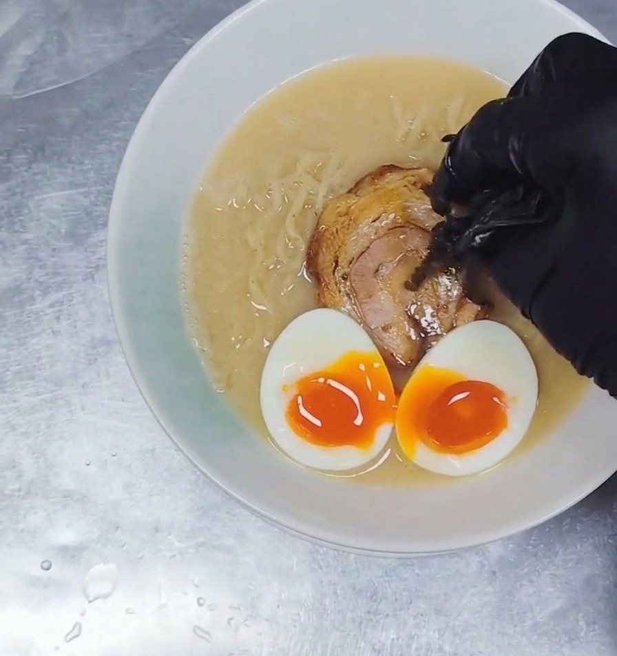 Dünne Streifen von dunklen Mu-Err-Pilzen werden zu einer Schüssel Ramen hinzugefügt, die mit Brühe, Nudeln, Fleisch und weichgekochten Eiern gefüllt ist.