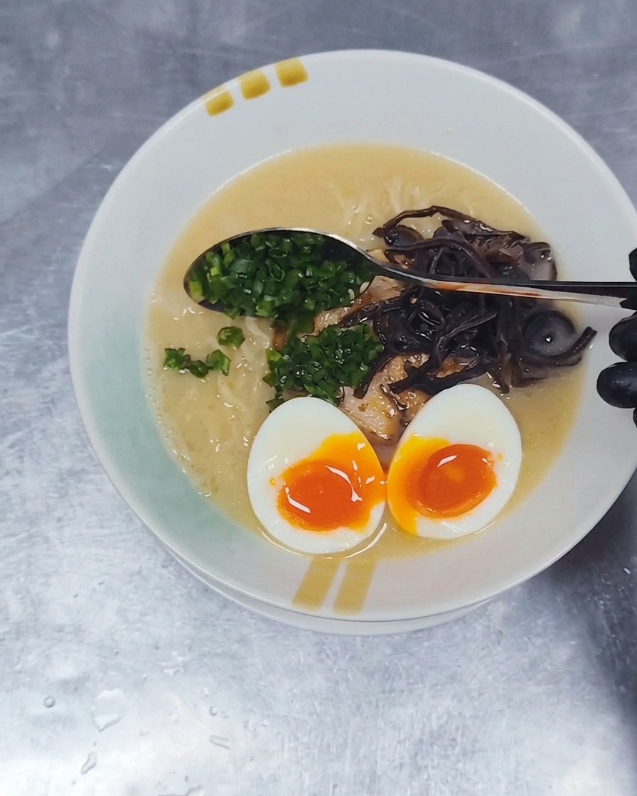Ein Metalllöffel streut fein gewürfelte grüne Frühlingszwiebeln auf die Oberfläche einer Tonkotsu-Ramenschüssel.
