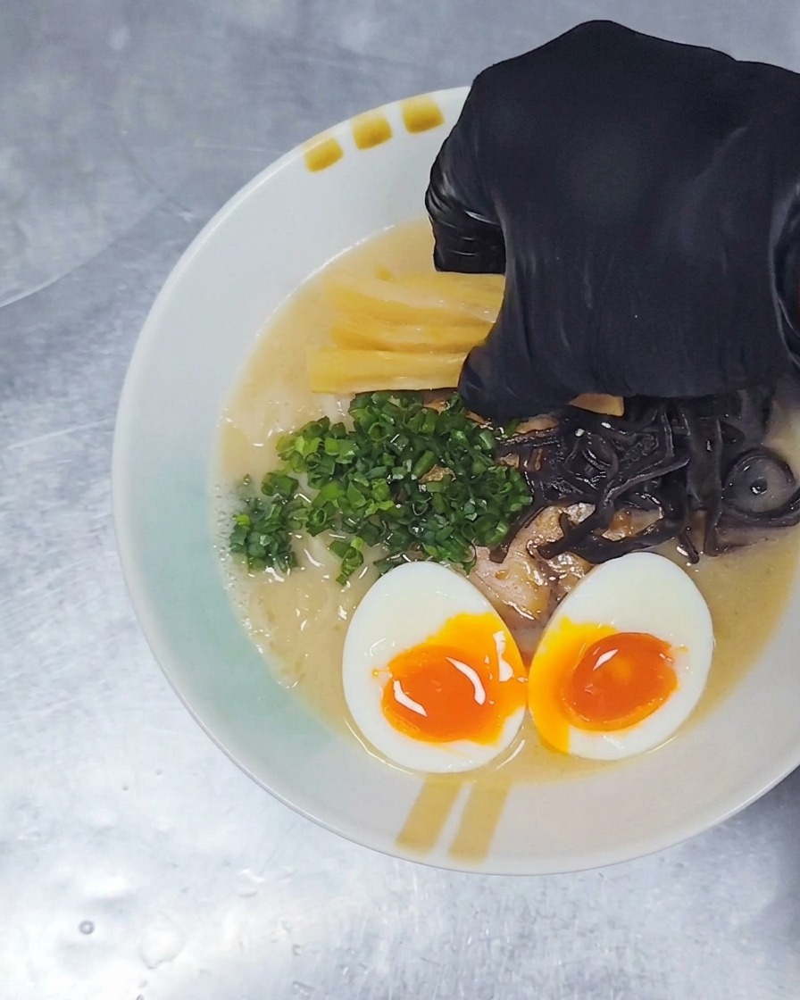 Eine Hand in einem Handschuh legt gelbe Streifen von gewürzten Bambussprossen auf eine Schüssel Ramen, die bereits mit Fleisch, Ei und Frühlingszwiebeln garniert ist.
