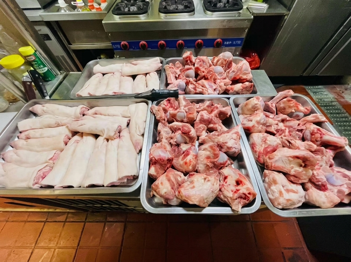Vier Metallbleche gefüllt mit rohen, rosa Schweineknochen und Schweinefüßen auf einer Küchentheke aus Edelstahl.