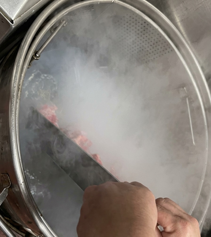 Hände halten ein großes Rührpaddel aus Metall und bewegen Schweineknochen in einem dampfenden Kochtopf.