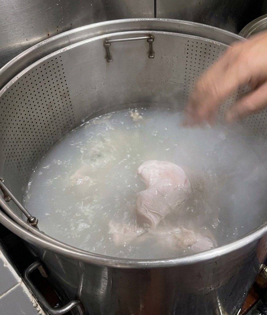Schweineknochenstücke in einem großen Topf mit kochendem Wasser, das sich von klar zu einem trüben, milchigen Weiß verfärbt.