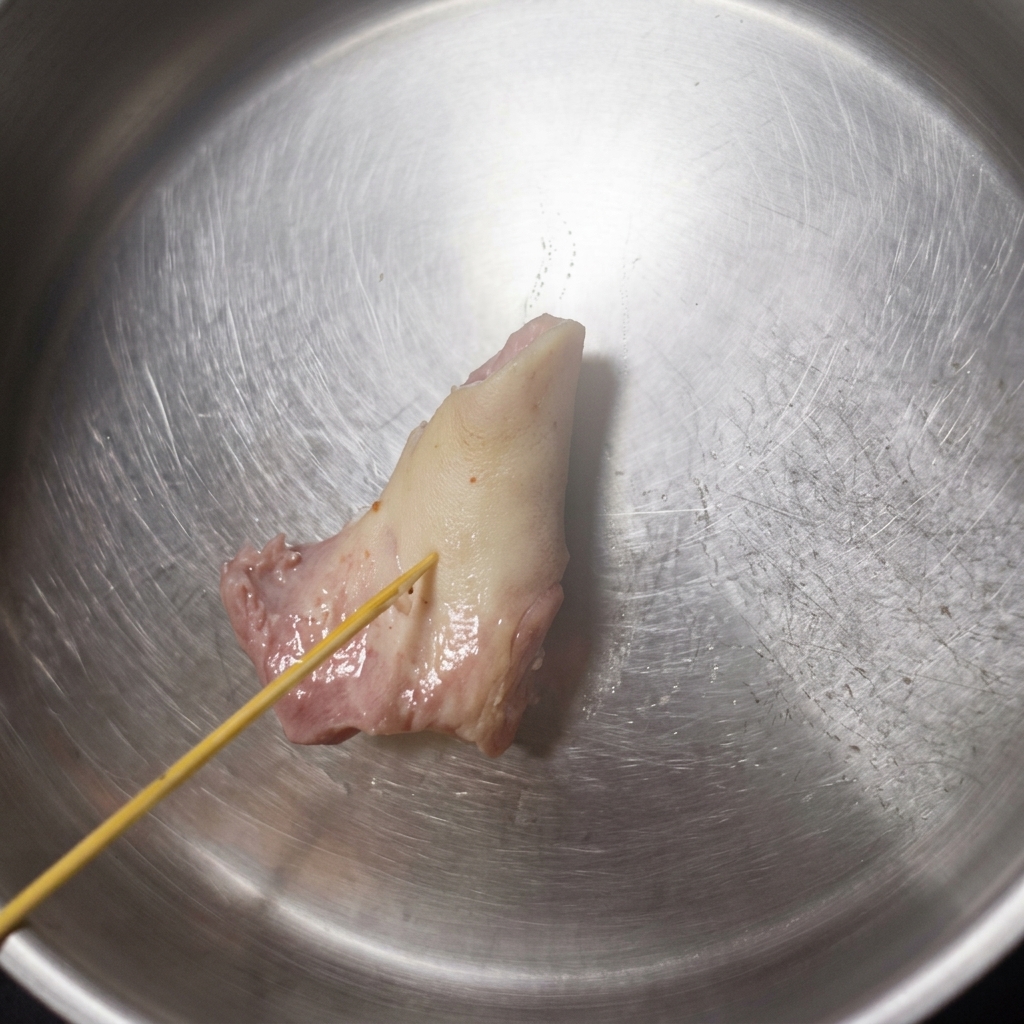 Ein Holzspieß drückt in einen hellen, aufgeweichten Schweineknochen in einer Edelstahlschüssel, wobei der Knochen nachgibt.