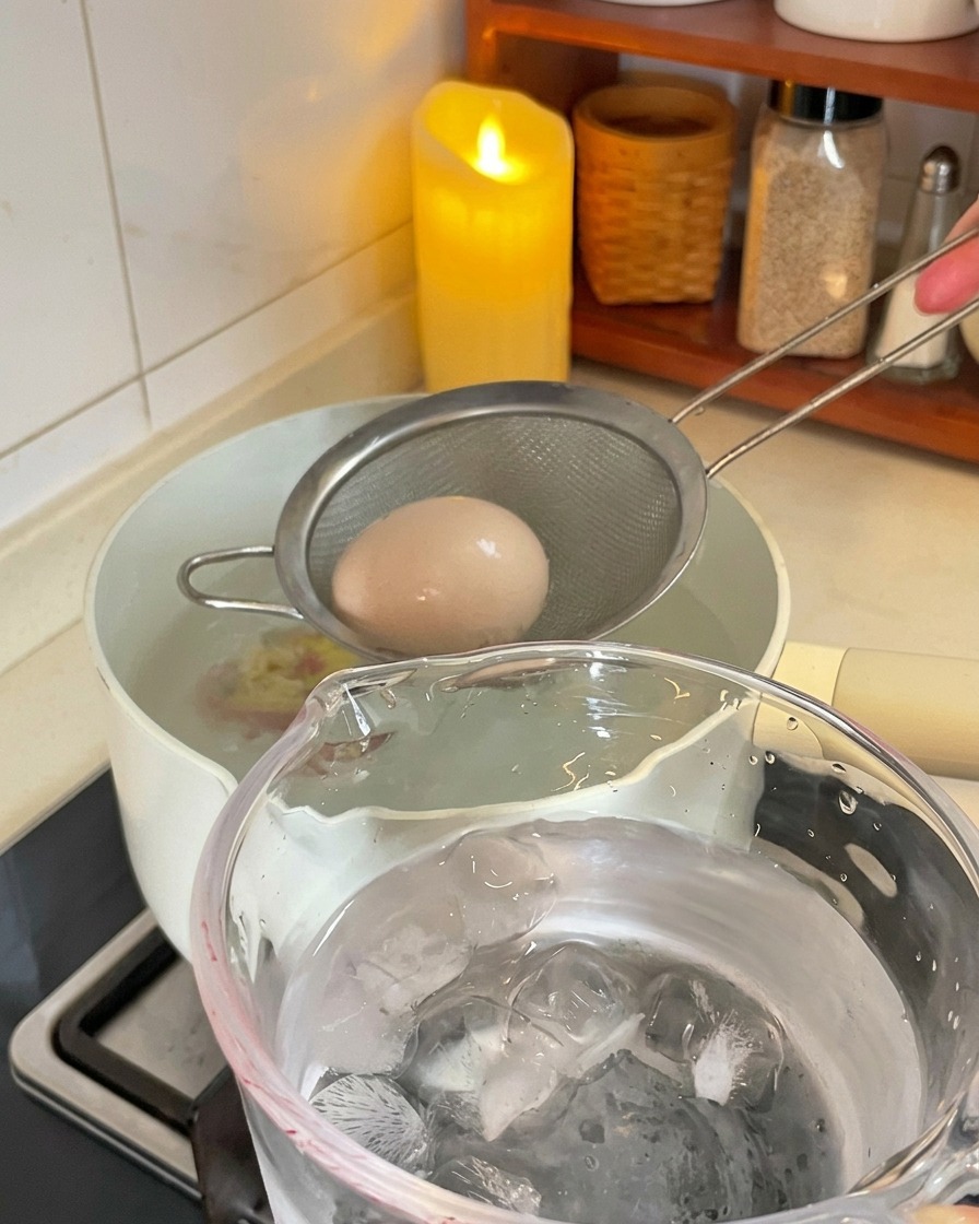 Ein kleiner Metallschaumlöffel hebt ein braunes gekochtes Ei aus einem weißen Topf und bewegt es in Richtung eines Glasbechers voller Eiswürfel und Wasser.
