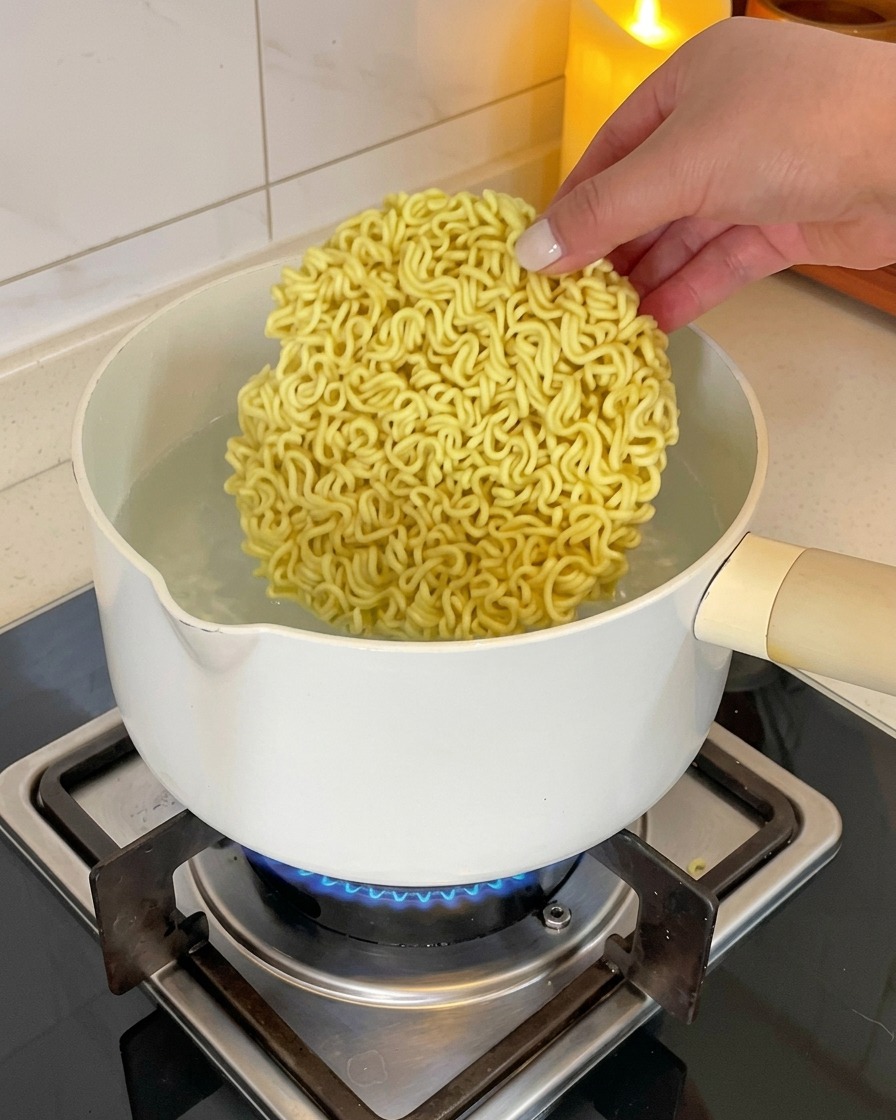 Eine Hand legt einen runden Block trockener, gewellter Instant-Ramen-Nudeln in einen weißen Topf mit kochendem Wasser auf einem Gasherd.