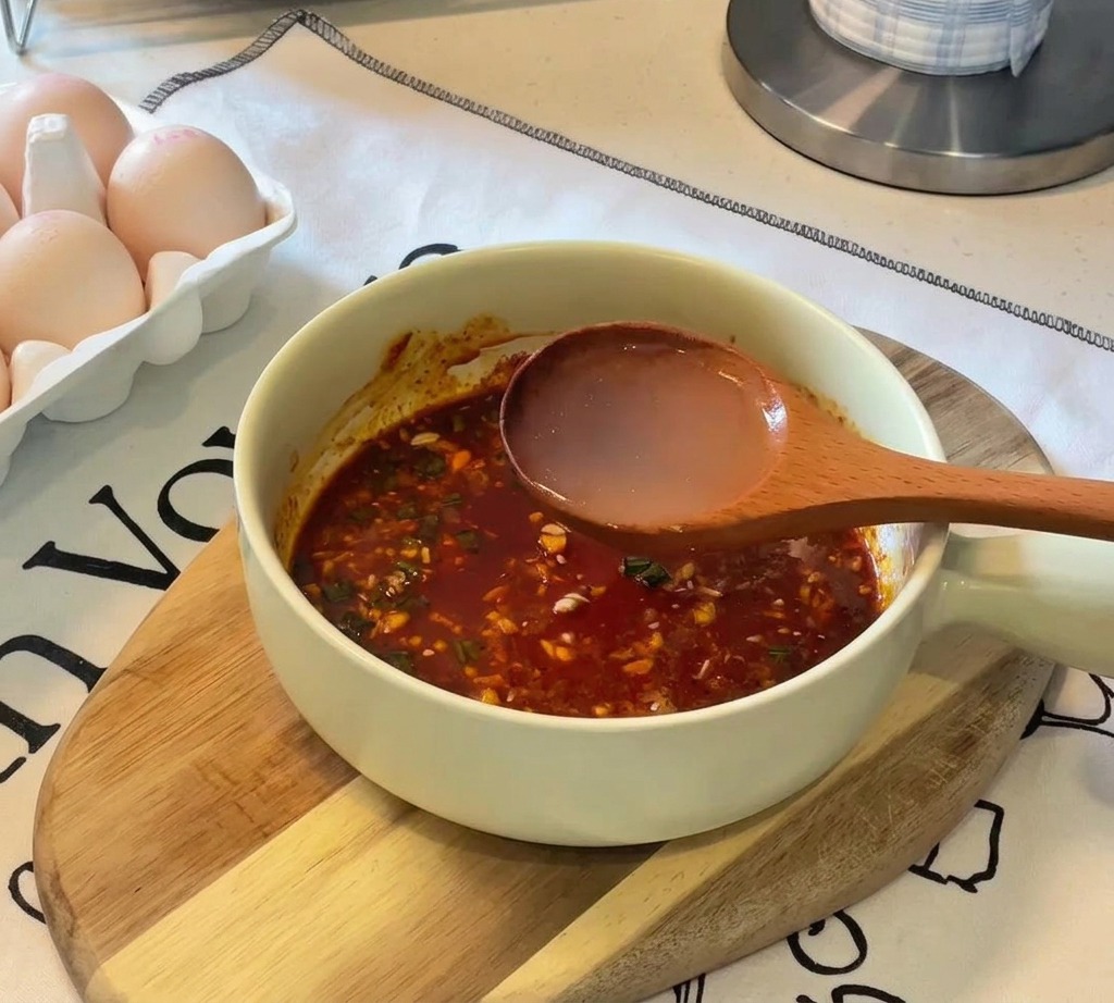 Eine Schöpfkelle aus Holz gießt klares, heißes Wasser in eine helle Schüssel, in der sich eine dunkle, rötlich-braune Gewürzpaste befindet.