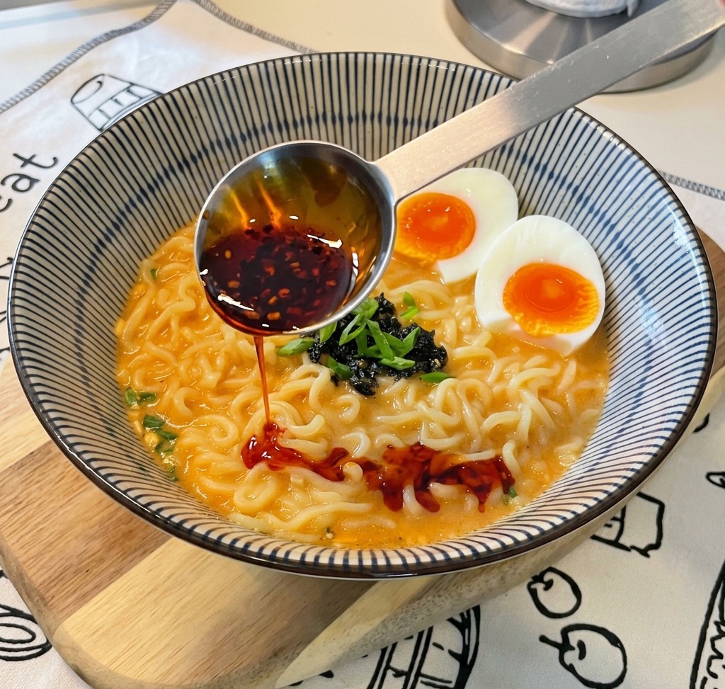 Ein Messlöffel aus Metall gießt leuchtend rotes Chiliöl mit sichtbaren Chiliflocken in eine Schüssel mit fertig zubereitetem Ramen.