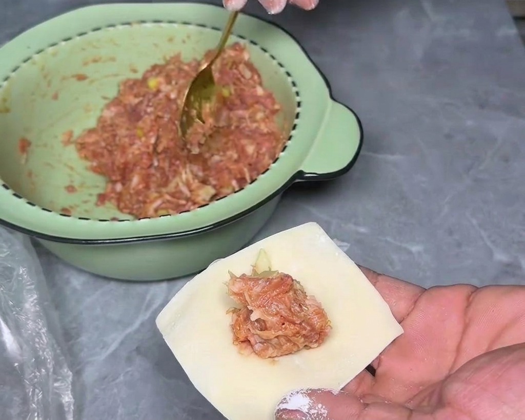 Eine Nahaufnahme einer Hand, die ein einzelnes quadratisches Wantan-Teigblatt mit einem kleinen Haufen Fleischfüllung in der Mitte hält.