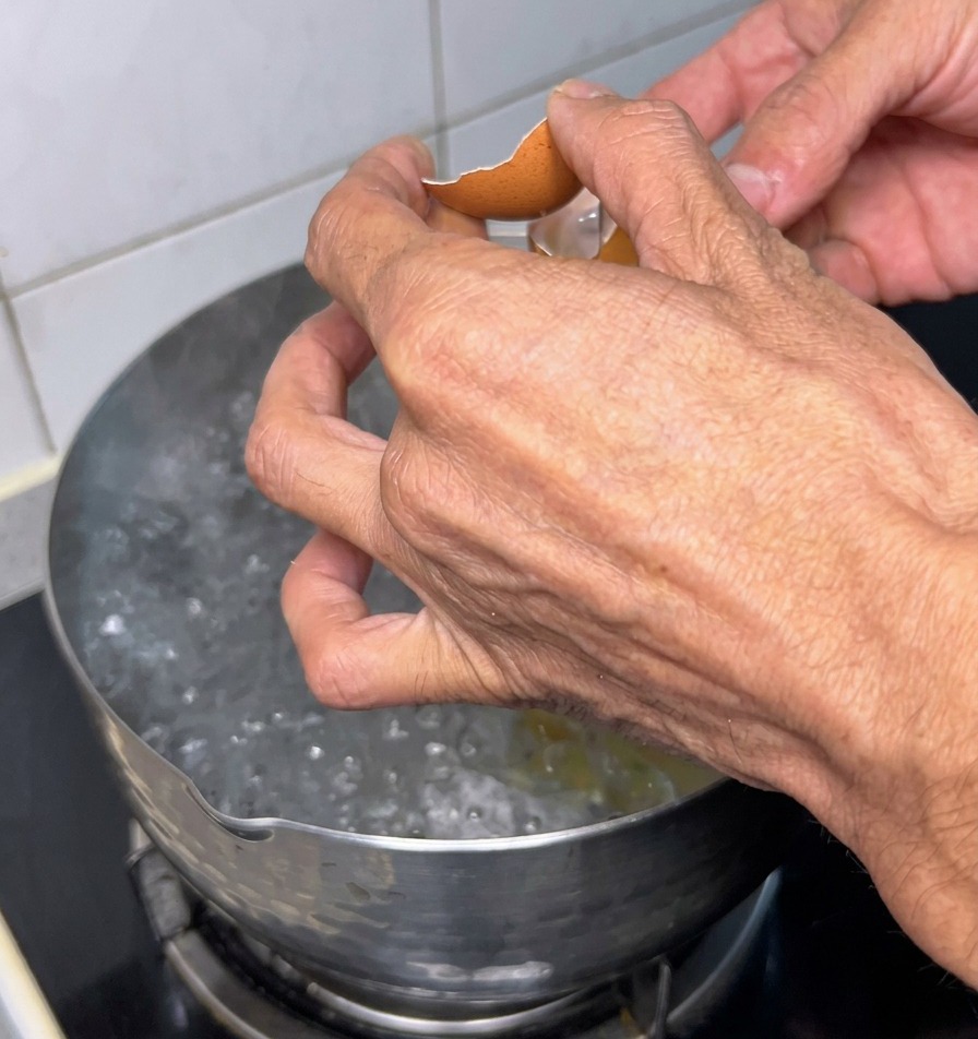 Zwei Hände schlagen die Schale eines braunen Eies über einem Edelstahltopf mit sprudelnd kochendem Wasser auf.