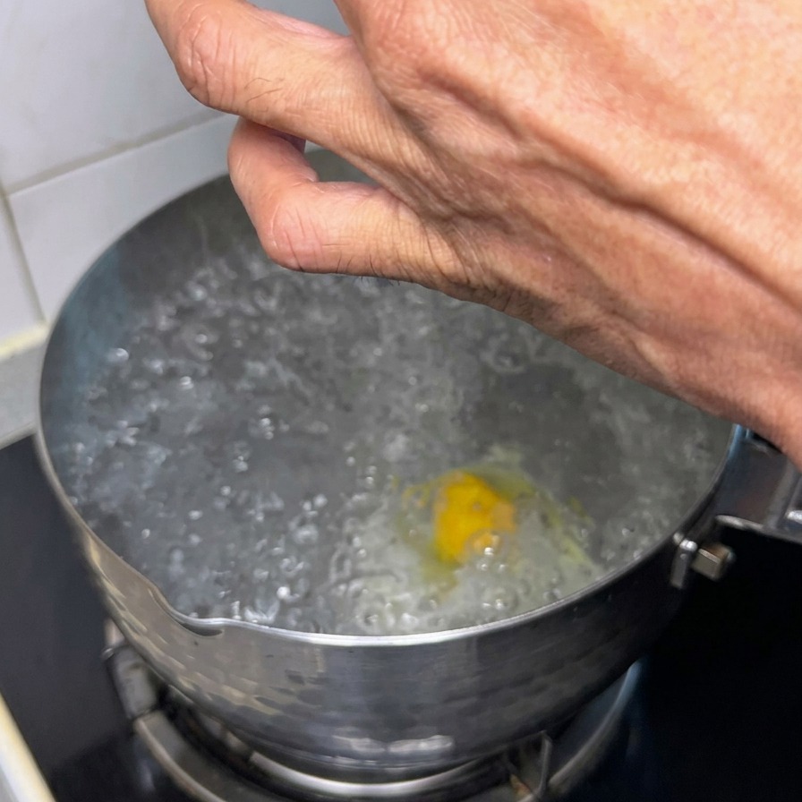 Eine Hand lässt mit einem Löffel eine kleine Portion rohes, gewürztes Fleisch in einen Topf mit kochendem Wasser gleiten, in dem bereits ein Ei pochiert.