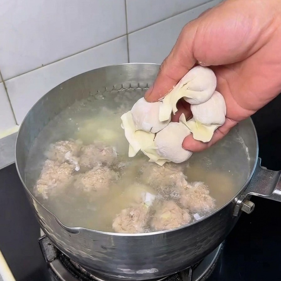 Die Hand einer Person gibt frische, rohe Wantans in einen Edelstahltopf mit kochendem Wasser, in dem sich bereits Fleischbällchen befinden.