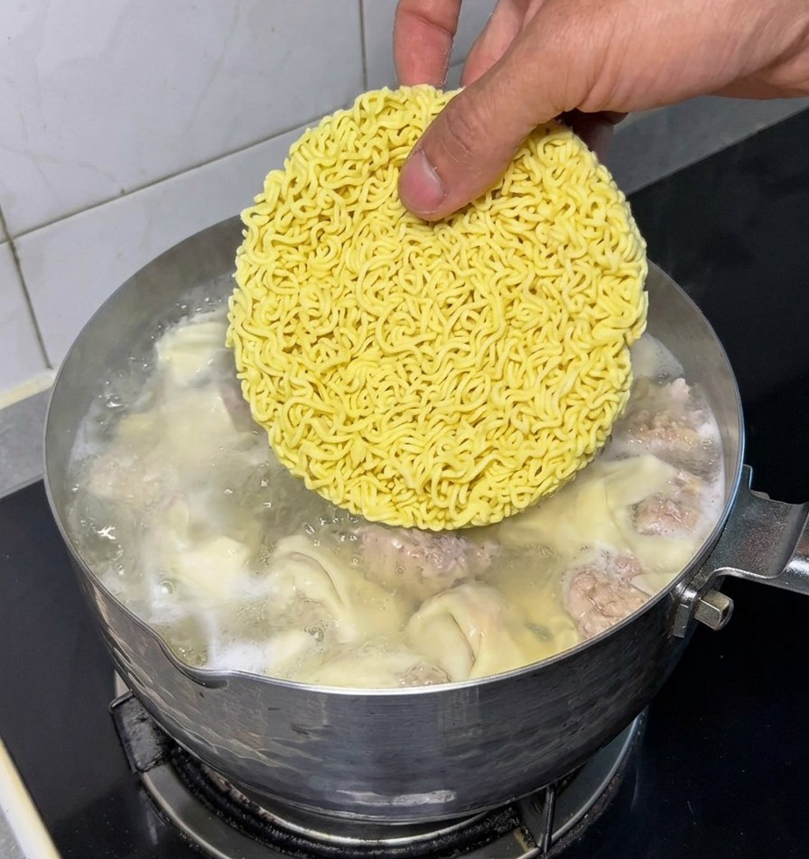 Eine Hand legt einen runden, gelben Block getrockneter Instant-Nudeln in einen Topf mit kochendem Wasser, der fertige Wantans und Fleischbällchen enthält.