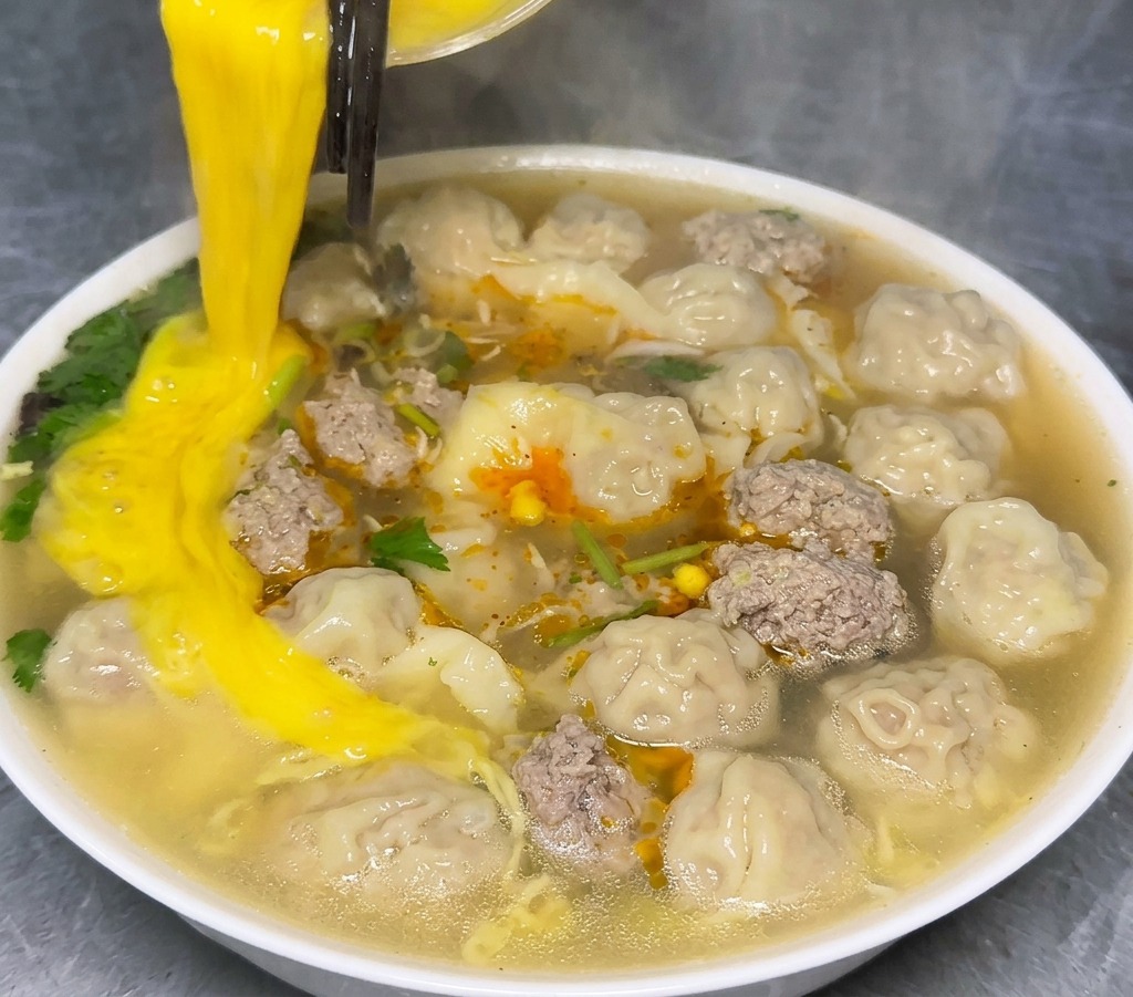 Verquirltes Ei wird aus einer Glasschüssel in eine weiße Keramikschüssel gegossen, die mit dampfender Wantan-Nudelsuppe gefüllt ist.
