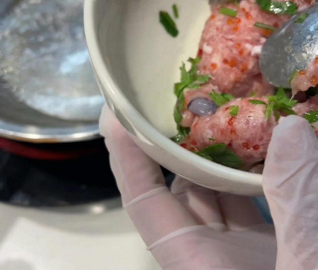 Eine behandschuhte Hand hält eine kleine weiße Schüssel mit rohem Schweinehackfleisch, leuchtend orangefarbenem Fischrogen, grünem Koriander und frischen Garnelen.