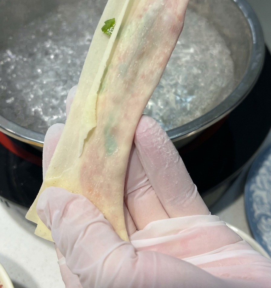 Behandschuhte Hände ziehen ein gefülltes Wan-Tan-Blatt über einem Topf mit kochendem Wasser in die Länge zu einem feinen Nudelband.