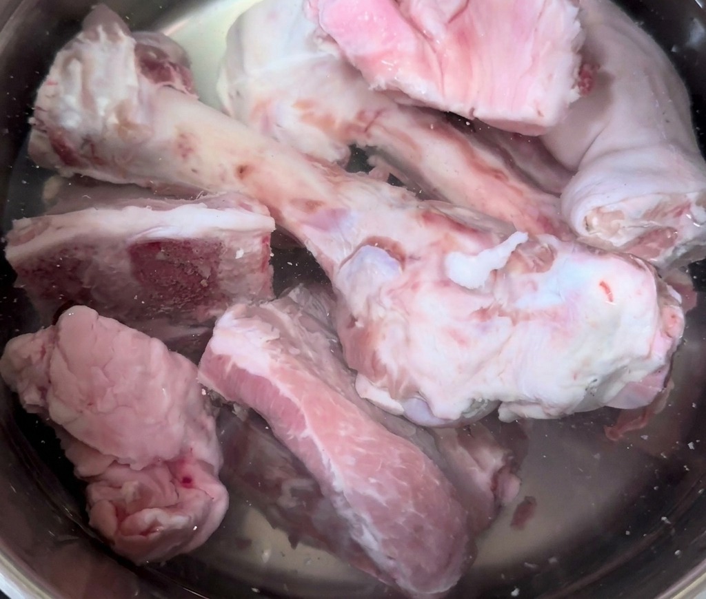 Rohe Schweineknochen und Schweinefüße in einem großen Edelstahltopf, der mit kaltem Wasser gefüllt wird.