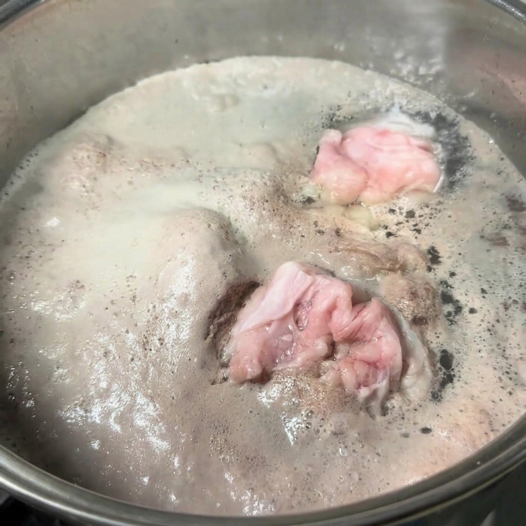 Ein Edelstahltopf mit kochendem Wasser und Schweineknochen, auf dessen Oberfläche eine dicke Schicht grauer Schaum schwimmt.
