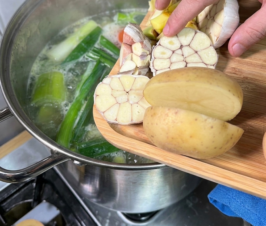 Eine Hand gibt halbierte Knoblauchknollen und Kartoffeln von einem Holzbrett in einen dampfenden Topf mit Knochenbrühe.