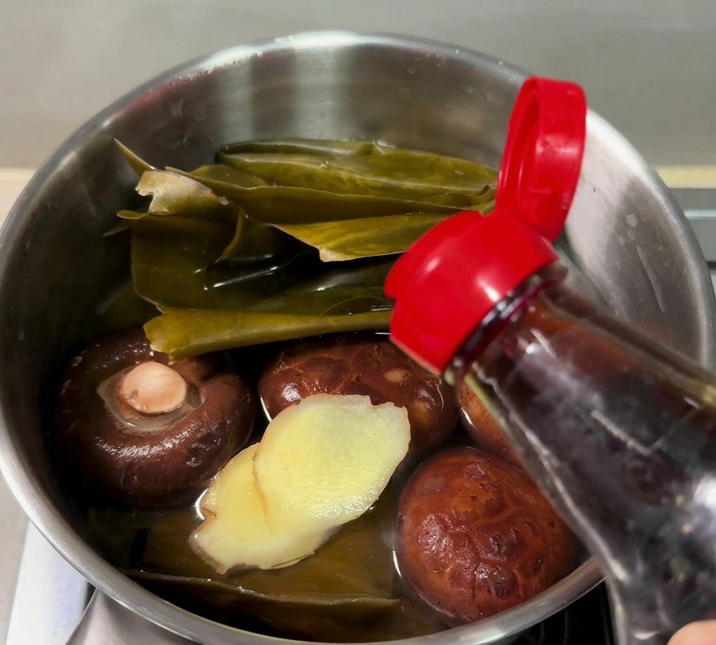 Fischsauce wird aus einer Flasche in einen kleinen Stieltopf mit Kombu, Shiitake-Pilzen und geschnittenem Ingwer gegossen.
