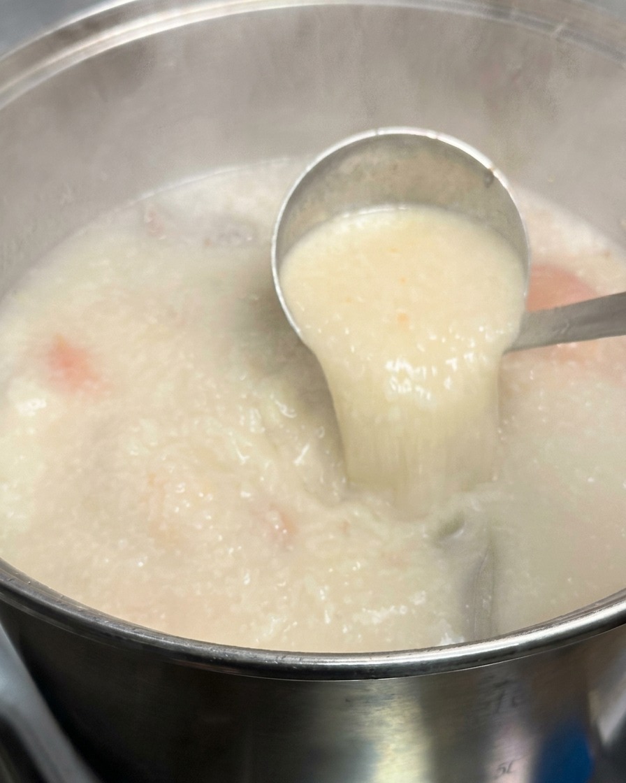 Eine Schöpfkelle aus Metall hebt eine Portion dicke, opake, milchig-weiße Tonkotsu-Brühe aus einem großen, köchelnden Suppentopf.