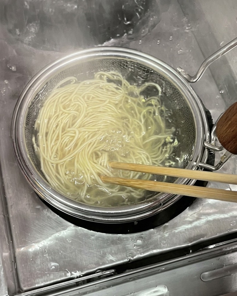 Ein Bündel dünner, blasser Weizennudeln wird mit Holzstäbchen in einem runden Nudelsieb aus kochendem Wasser gehoben.