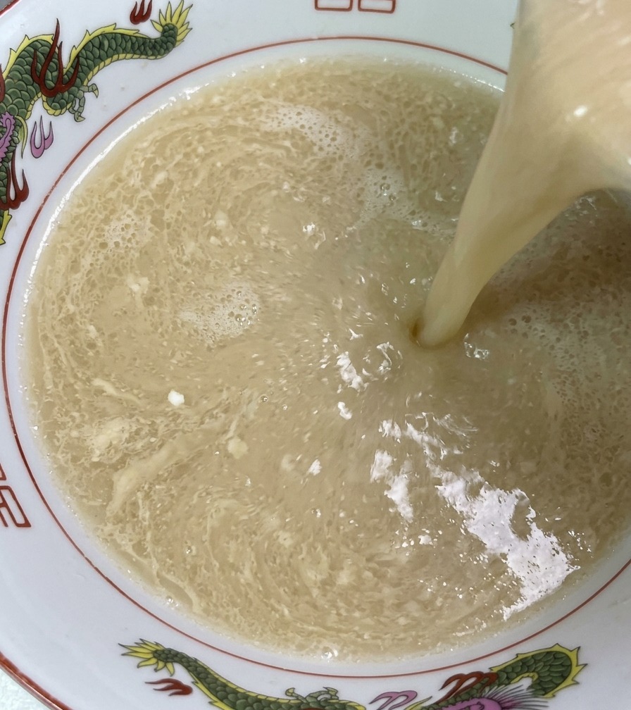 Dampfende, elfenbeinfarbene Schweineknochenbrühe wird aus einer Kelle in eine dekorierte Ramenschüssel gegossen, wodurch die reichhaltige und seidige Textur sichtbar wird.