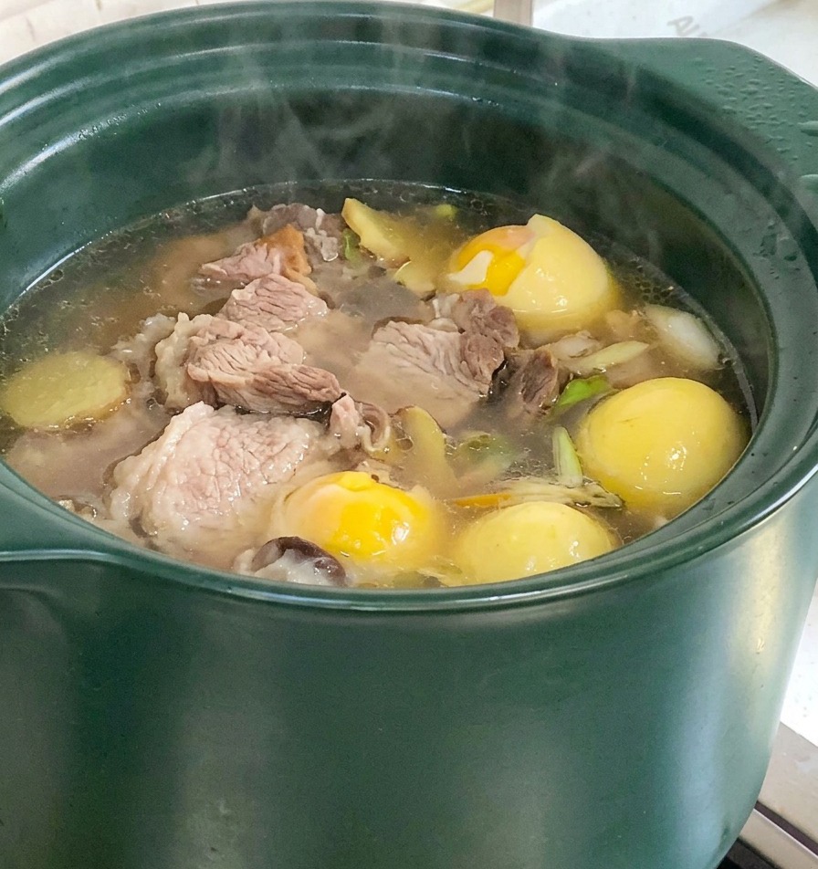 Ein grüner Tontopf mit köchelnder Schweineknochensuppe, in der Maisstücke und Yams-Wurzeln zu sehen sind.