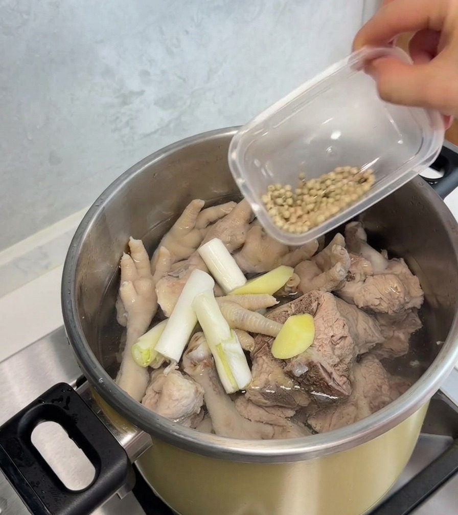 Eine Hand gibt weiße Pfefferkörner aus einem kleinen Plastikbehälter in einen Edelstahltopf mit blanchierten Hühnerfüßen, Schweineknochen, Frühlingszwiebeln und Ingwerscheiben.