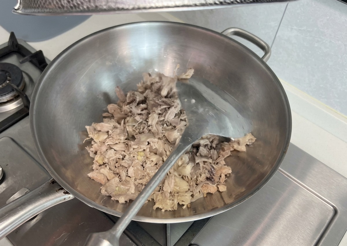 Zerkleinerte, gekochte Fleischreste werden in einem großen Edelstahl-Wok auf dem Herd angebraten und mit einem Metallwender zerdrückt.
