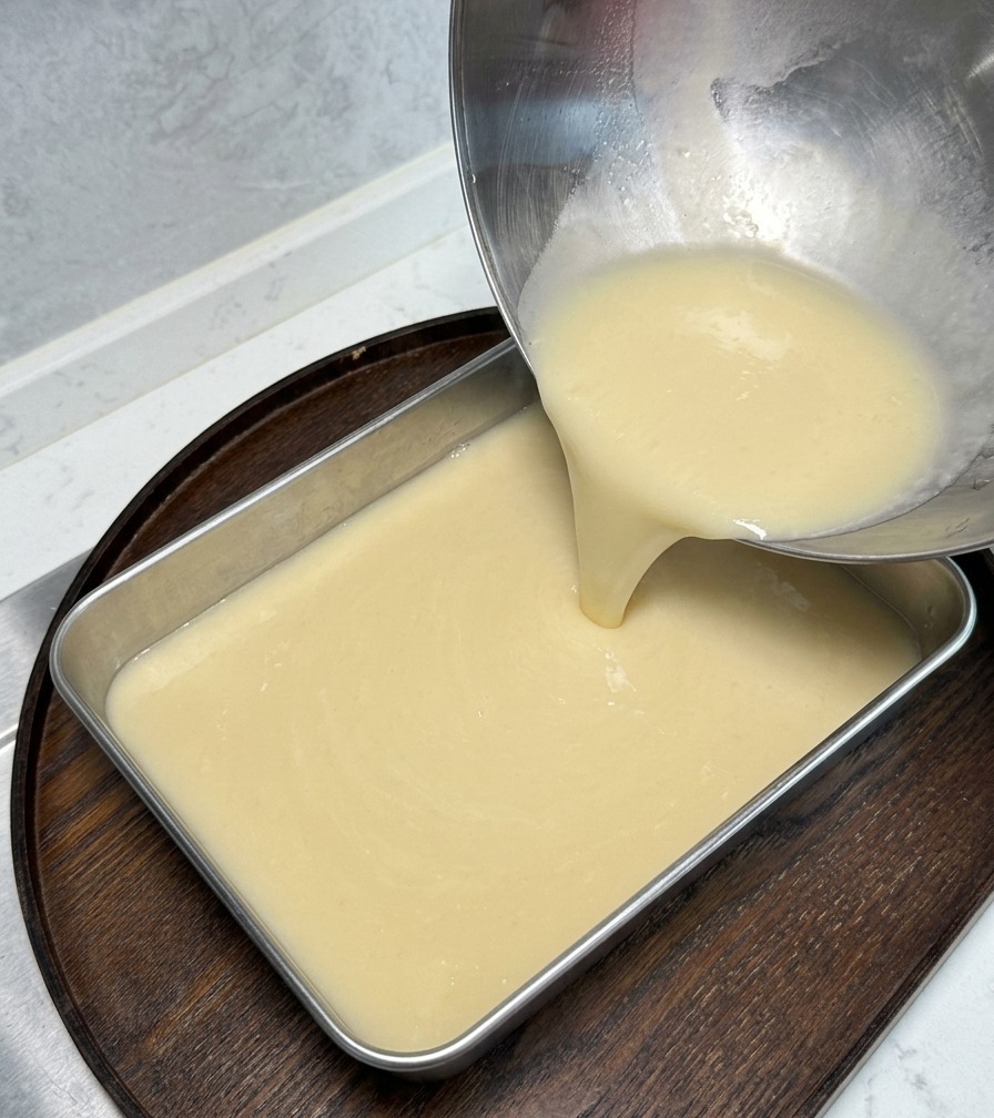 Eine dicke, cremefarbene Brühe wird aus einer Edelstahlschale in eine rechteckige Metallform auf einem Holzbrett gegossen.