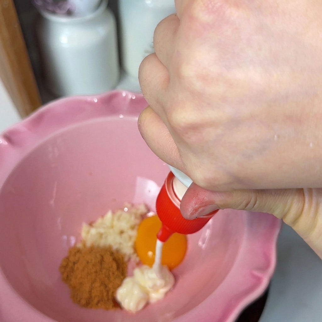 Weiße Mayonnaise wird aus einer roten Tülle in eine rosafarbene, geriffelte Schüssel gedrückt, die gehackten Knoblauch, ein leuchtend orangefarbenes Eigelb und braunes Gewürzpulver enthält.