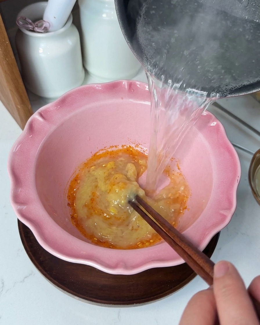 Heiße, klare Flüssigkeit wird aus einem Topf in eine rosafarbene Schüssel mit einer cremigen, gelben Gewürzpaste gegossen und mit Holzstäbchen umgerührt.