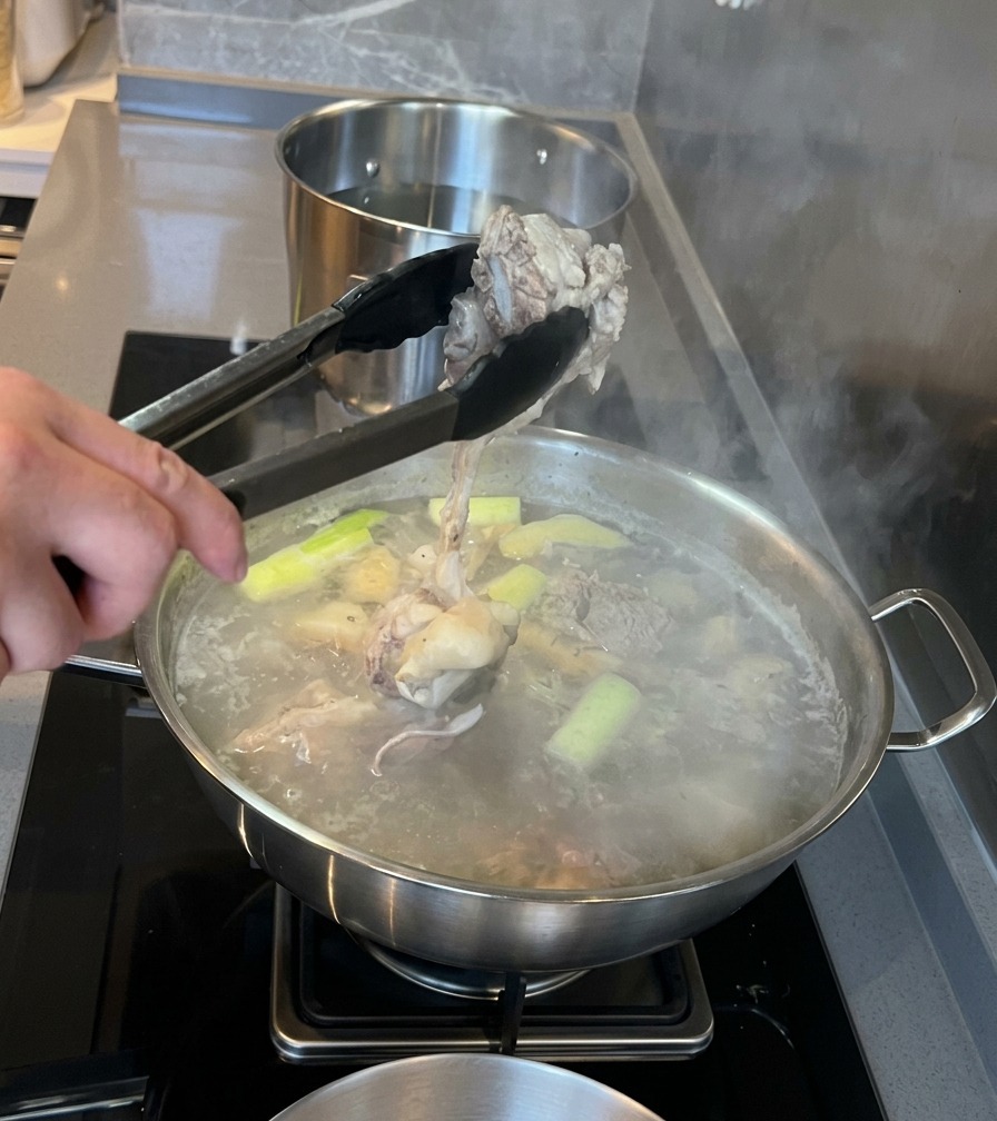 Eine Küchenzange hebt einen blanchierten Schweinemarkknochen aus einem Topf mit köchelndem Wasser und Gewürzen.