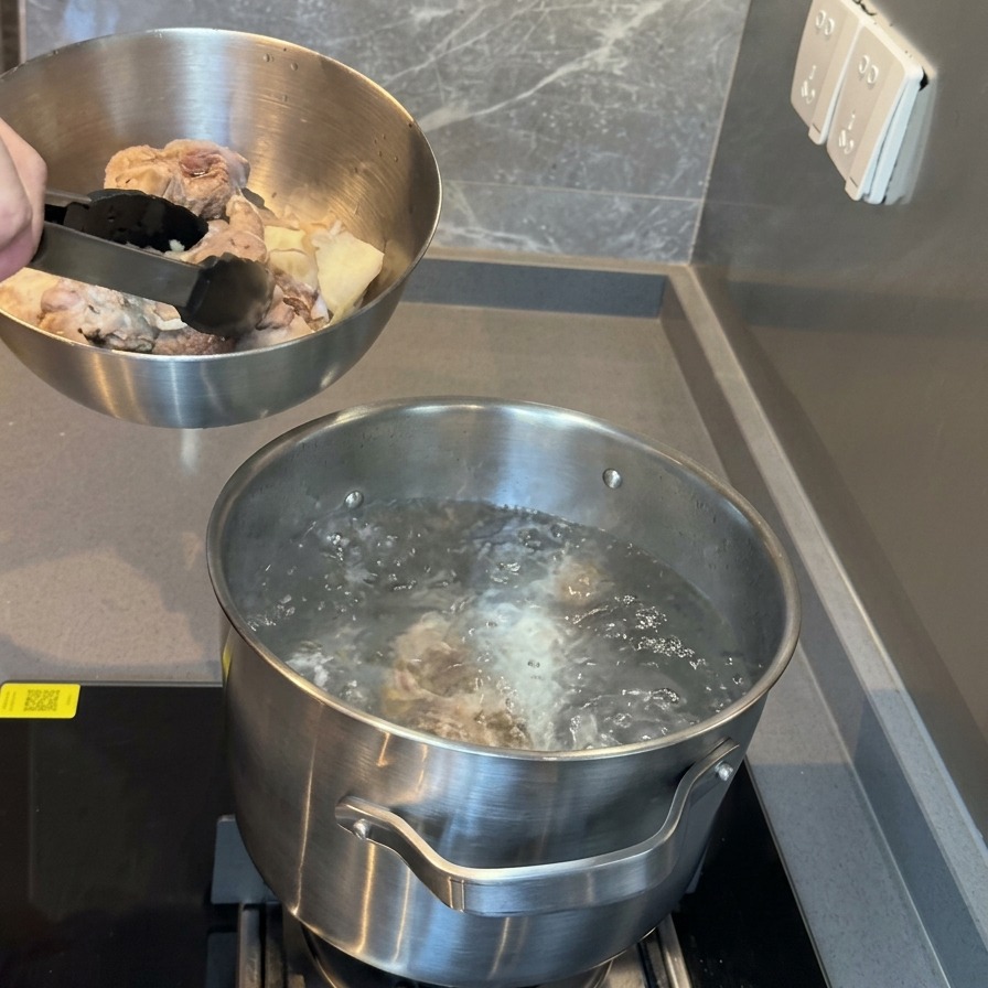 Eine Metallschüssel mit blanchierten Schweineknochen wird über einen Topf mit kochendem Wasser gehalten, bereit zum Umfüllen.