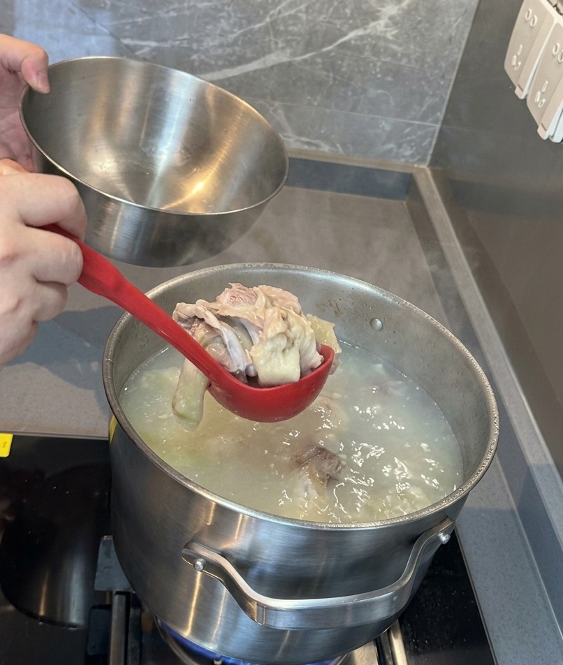 Eine rote Schöpfkelle hebt weiche Schweineknochen und Fleisch aus einer milchig-weißen Brühe in eine Edelstahlschüssel.