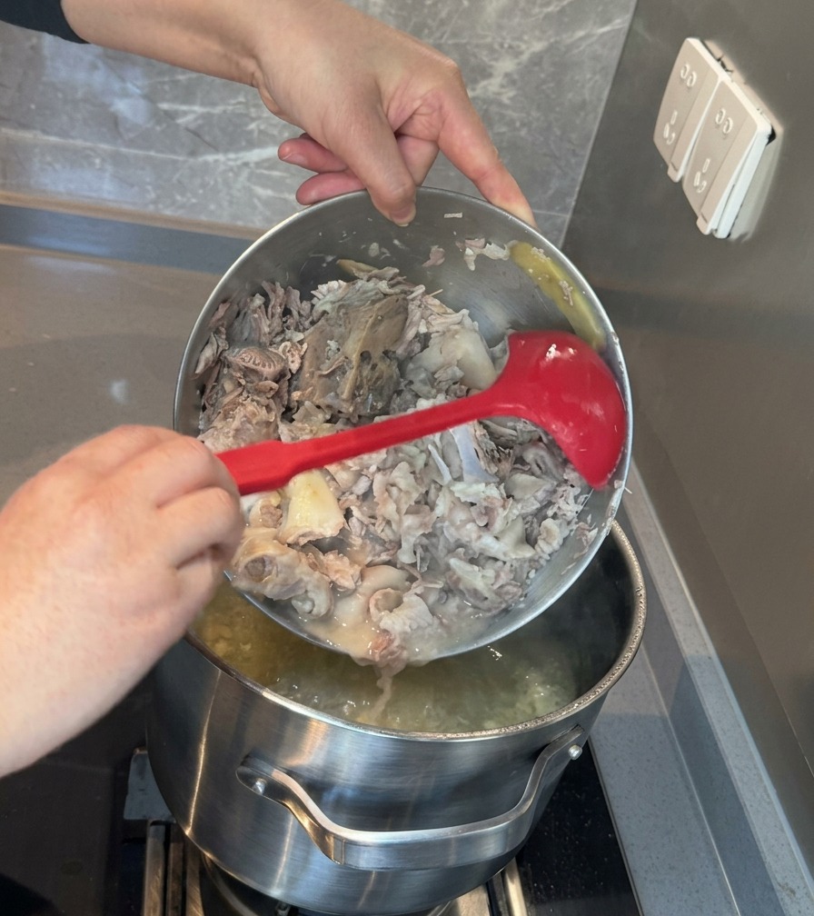 Der Inhalt einer Metallschüssel mit zerdrücktem Fleisch und Knochen wird zurück in den großen Topf mit weißer Brühe geschüttet.