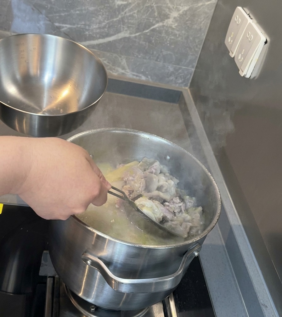 Ein feines Edelstahlsieb filtert Knochen- und Fleischreste aus einem tiefen Topf mit milchig-weißer Schweinesuppe.
