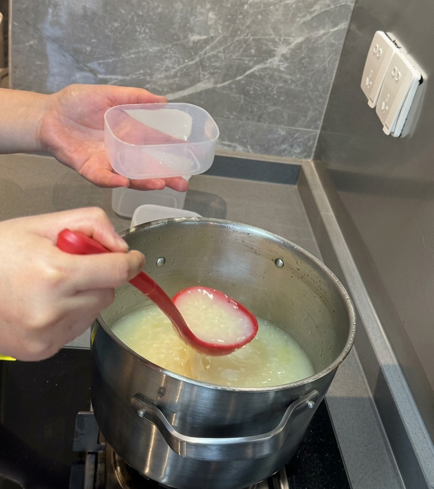 Eine rote Schöpfkelle gießt die weiße Schweinebrühe in einen kleinen, durchsichtigen Vorratsbehälter aus Plastik.