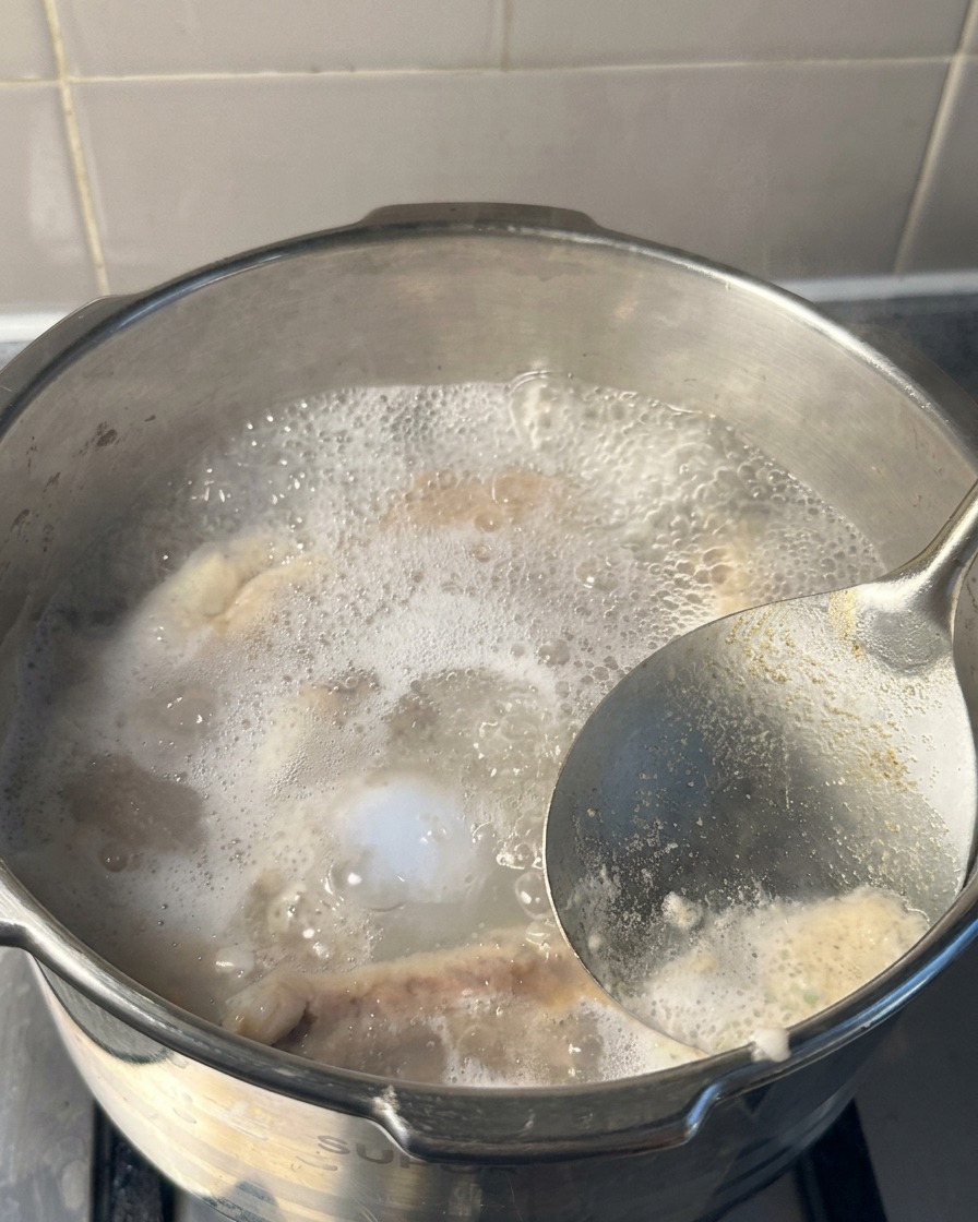 Eine Nahaufnahme einer Metallkelle, die weißen und grauen Schaum von der Oberfläche des kochenden Wassers in einem Schnellkochtopf abschöpft.
