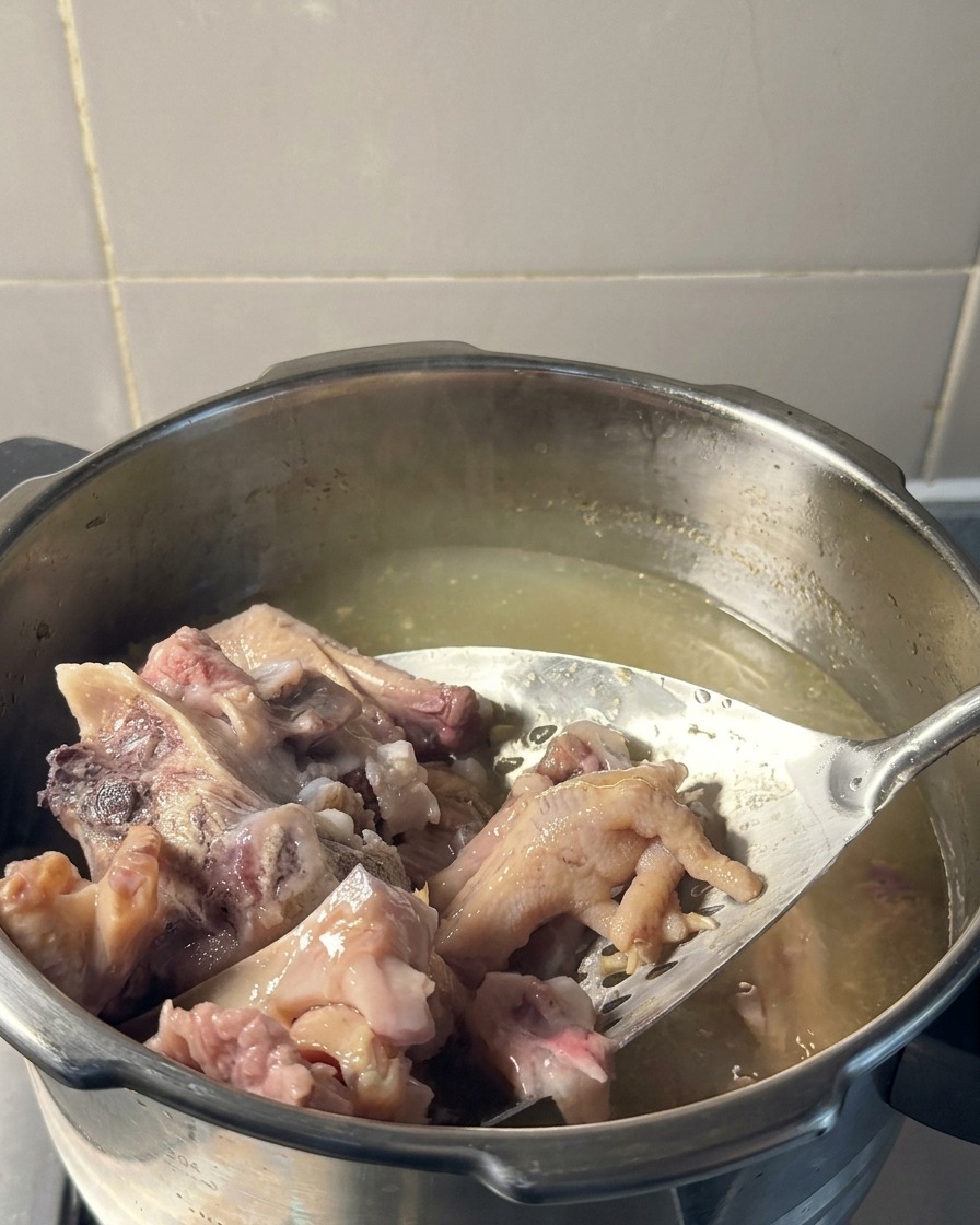 Ein Schaumlöffel hebt zartes, gekochtes Schweinefleisch und Hühnerfüße aus einem Topf mit blassgelber Brühe.