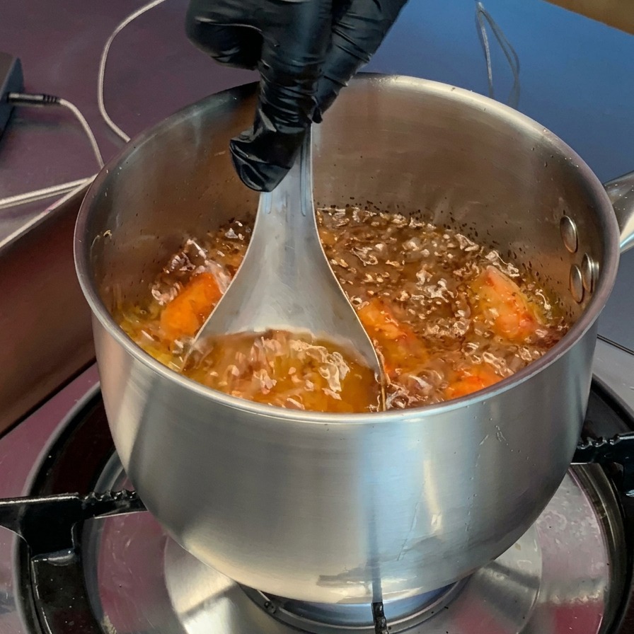 Eine Hand mit Handschuh drückt orangefarbene, gegarte Garnelenköpfe mit einer Schöpfkelle aus Metall in das heiße Öl im Topf.