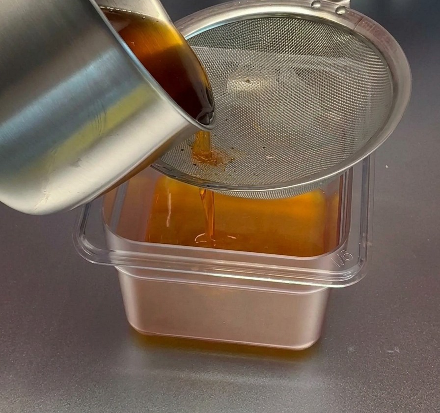 Rot-orangefarbenes Garnelenöl wird aus einem Edelstahltopf durch ein feines Metallsieb in einen klaren, rechteckigen Behälter gegossen.