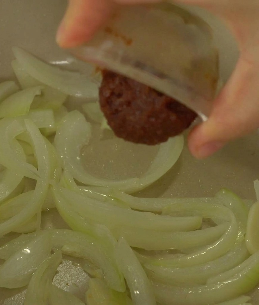 Eine Hand gibt aus einer kleinen Schüssel einen Klecks dunkelbraune fermentierte Sojabohnenpaste zu den glasigen Zwiebeln im Topf.