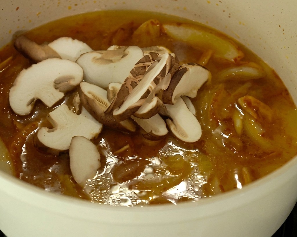 Frisch geschnittene Pilze und Kohlstücke schwimmen auf der Oberfläche einer kräftig orangefarbenen, leicht köchelnden Ramen-Brühe in einem weißen Topf.