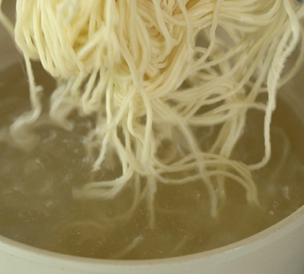 Ein Bündel dünner, heller Ramen-Nudeln wird in einen Topf mit sprudelnd kochendem Wasser gegeben.