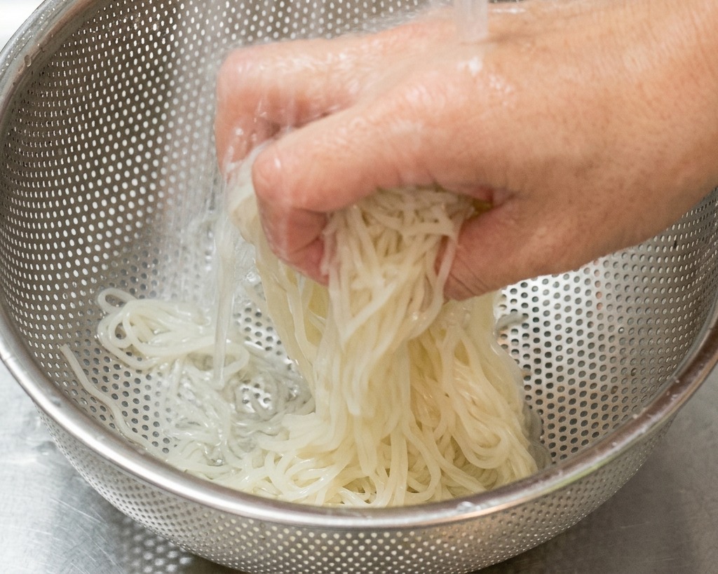 Eine Hand schwenkt helle Ramen-Nudeln in einem Edelstahlsieb unter kaltem fließendem Wasser im Spülbecken.