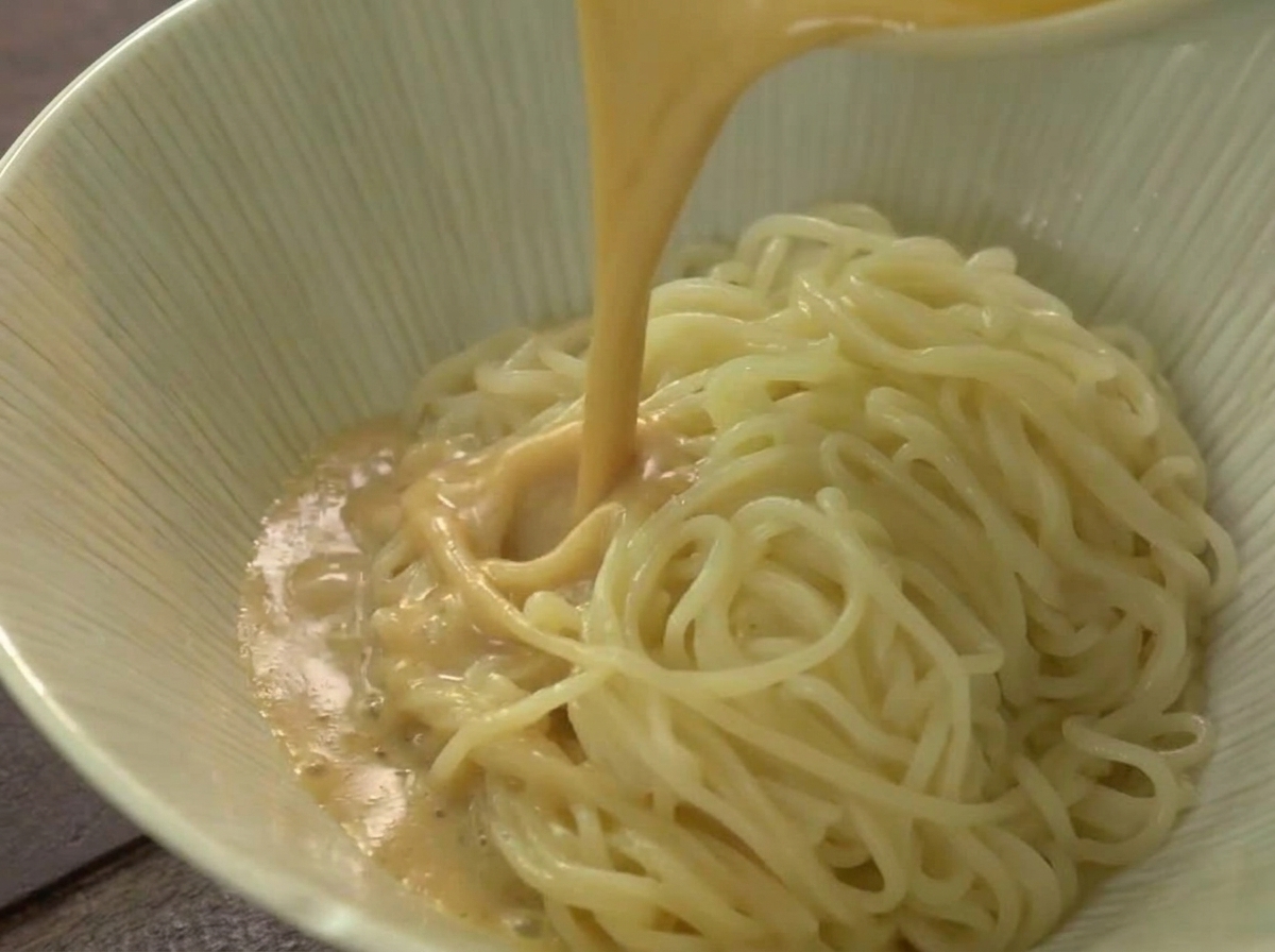 Ein Strahl heißer, cremefarbener Ramen-Brühe wird über einen Berg Nudeln in einer hellen Keramikschüssel gegossen.