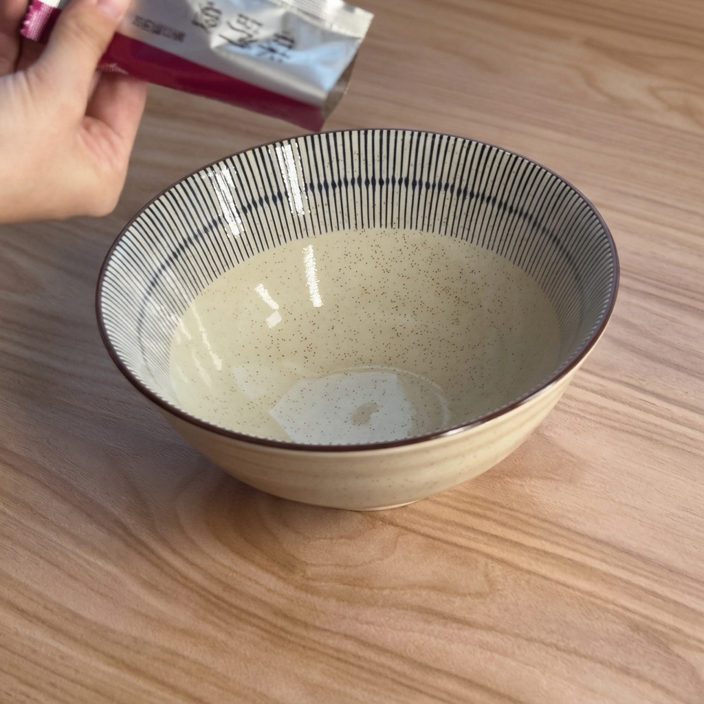 Eine Hand drückt braune Miso-Paste aus einer silbernen Folienpackung in eine cremefarbene Keramikschüssel mit blauen Streifen.